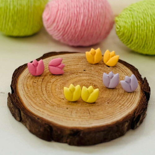 Tulip Stud Earrings in pastel pink, yellow, purple, and orange, arranged on a wooden slice with spring-colored yarn in the background.